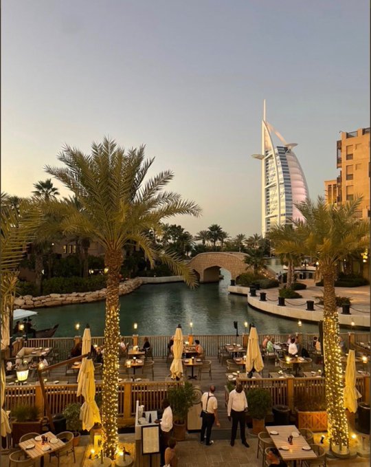 The image shows an outdoor restaurant patio with several tables and chairs set up for guests. The patio overlooks a body of water, which features palm trees nearby. In the background, there are buildings that include a large hotel or resort structure. A group of people can be seen enjoying their time at the restaurant, while others are walking around in the area.