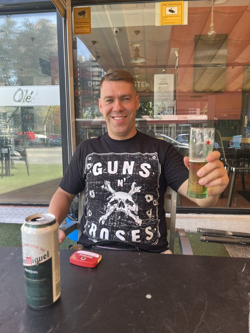 A man wearing a black Guns N' Roses t-shirt smiles while holding a glass of beer; a can of San Miguel beer sits on the table in front of him, alongside a red tin.