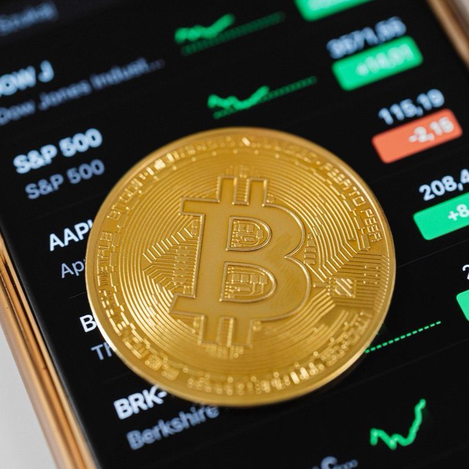 A gold Bitcoin token rests on a smartphone displaying a stock market app with tickers including S&P 500, AAPL, BRK (Berkshire), and green/red price indicators, visually representing the convergence of traditional equities and cryptocurrency markets relevant to Russia's proposal to allow Bitcoin, Ether, and USDT trading on regulated exchanges.
