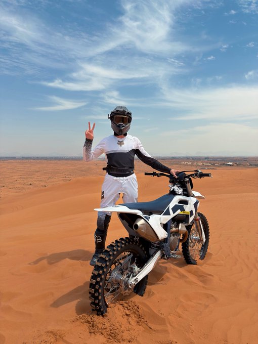 A person in a white and black motocross outfit and helmet posing with a peace sign next to a dirt bike in a desert landscape.