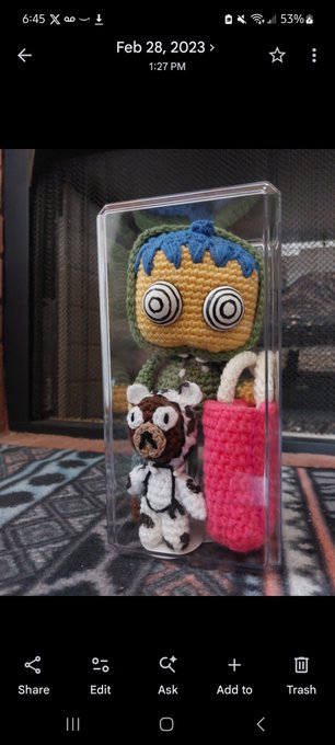 A crocheted amigurumi figure with blue "hair" and hypnotic eyes displayed in a clear protective case alongside a small bear plush and pink stitched accessory, photographed on February 28, 2023. The image appears to show a handmade craft item that the poster had promised to mail but failed to deliver, tagging recipients @berkinbags, @gettles, and @0xRadi0.