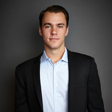 A professional headshot of a young man with short brown hair wearing a dark suit jacket over a light blue button-up shirt, posed against a dark gray background.
