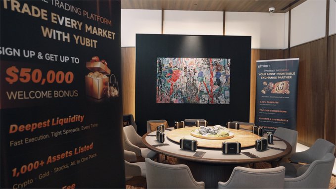 A private dining room or VIP meeting space with a large round wooden table surrounded by gray chairs, featuring Yubit promotional banners advertising a $50,000 welcome bonus, deepest liquidity, and 1,000+ assets listed, with a colorful painting on the wall.