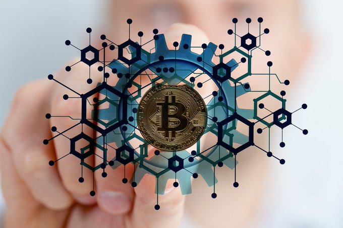 A conceptual image of a hand holding a Bitcoin coin in front of a digital blue gear design overlaid with blockchain network graphics.