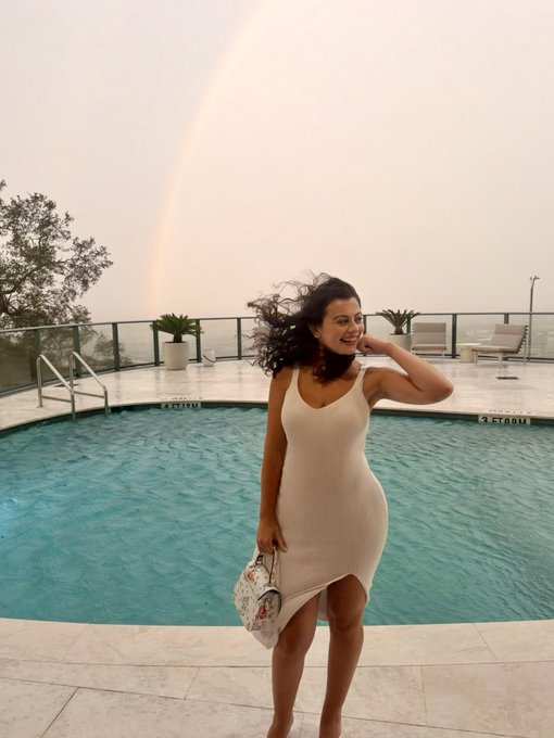 Photo of a woman in a cream-colored dress standing by a swimming pool with a rainbow visible in the misty sky behind her.