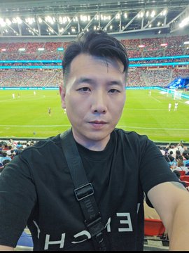 A selfie of a man wearing a black t-shirt with a camera strap, taken in what appears to be a sports stadium with a green field and stands in the background.