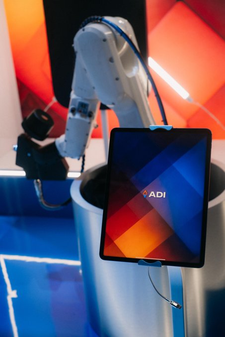 The image features a booth at a tech conference with a tablet computer displaying the word "ADI" on its screen. The booth has a robot arm attached, which appears to be controlling or interacting with the tablet. The scene suggests that the booth is promoting their product and showcasing it in an engaging manner.