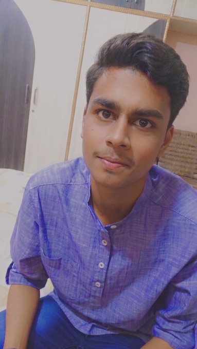 A young man with dark hair and a slight smile takes a selfie in a room with wooden cabinetry in the background, wearing a blue button-up kurta—a potential new profile picture to greet his followers with a warm "GM Everyone."