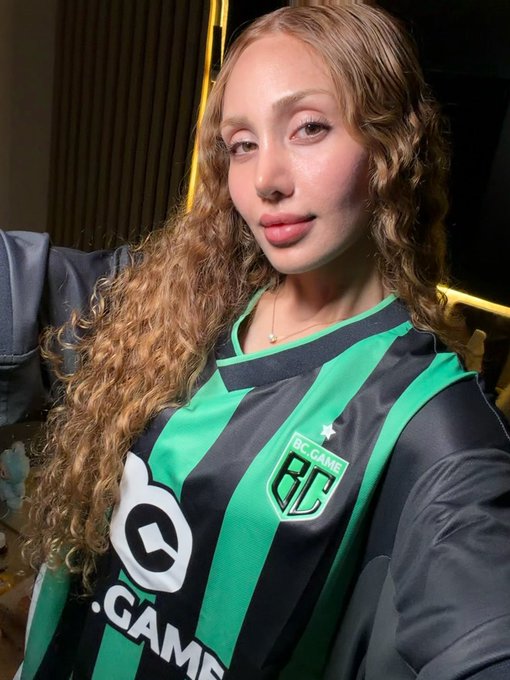 Selfie of a young woman with long curly hair wearing a green and black striped BC.Game jersey with a 'BC GAME' crest and '.GAME' sponsor logo.