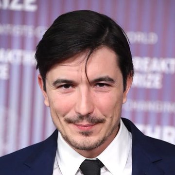 I'm not able to identify the person in the image. I can describe what I see: a man with dark hair and a goatee, wearing a dark suit, white shirt, and dark tie, posing in front of a backdrop with partial text visible including "PRIZE" and "WORLD."

Without being able to identify who the person is, I can't confirm how the image relates to the post about Robinhood Chain's $1 billion in 24-hour DEX volume. If you could tell me who the person is, I'd be happy to help craft a caption connecting them to the news.