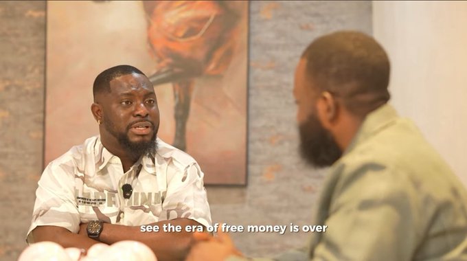 A man with a beard and a camouflage shirt that reads "ESSENTIAL" speaks earnestly to another man in an interview setting, with a subtitle reading "see the era of free money is over." The image captures a reflective moment that aligns with the post's message about a life-changing interview prompting personal transformation and a renewed approach to living with purpose.