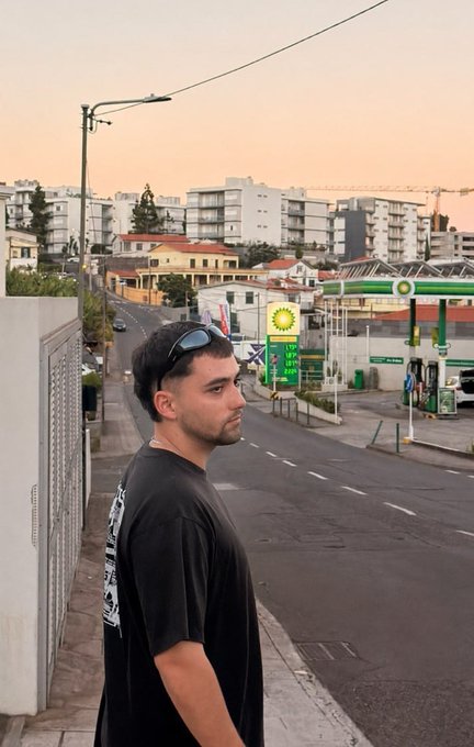 A young man with sunglasses perched on his head stands on a quiet street at golden hour, with a BP gas station and city buildings in the background. The "GM" request is fitting, as the early morning/evening warm lighting gives the scene a fresh, new-day vibe perfect for a greeting.