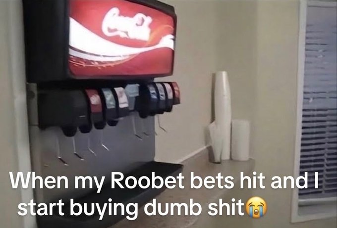 A Coca-Cola Freestyle soda fountain machine with a row of flavor dispensers stands against a wall next to stacked cups and window blinds. Caption text reads "When my Roobet bets hit and I start buying dumb shit 😭" — humorously suggesting that gambling winnings are being splurged on a luxury soda fountain, tying into the post's joke about peak financial freedom from Roobet winnings.