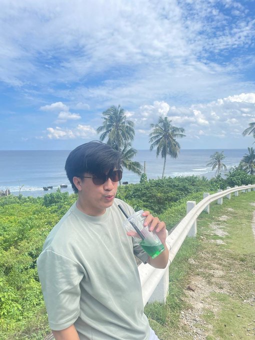 A young man in sunglasses sips a green drink while standing by a tropical oceanfront with palm trees and a coastal path in the background. The casual, vacation-style vibe complements the post's celebratory tone, highlighting the winner enjoying a $100 CAD bonus buy giveaway from @PP_Privatejet, selected by @SteveOnStake.