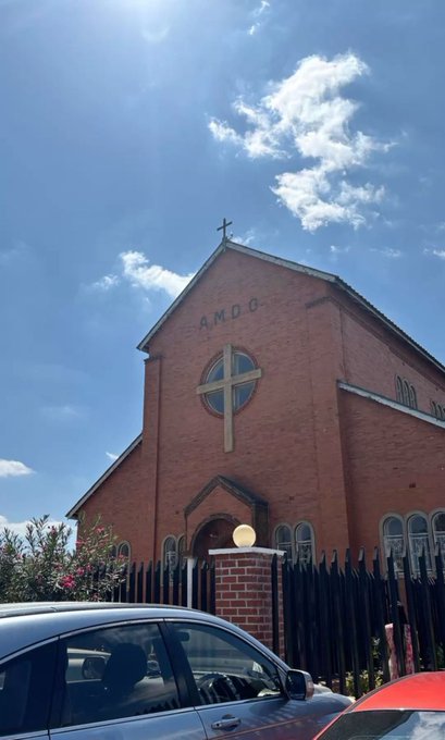 A red-brick church featuring "A.M.D.G." (Ad Majorem Dei Gloriam, meaning "For the Greater Glory of God") inscribed above a cross-topped gable, with a black car parked in front under a bright blue sky—fittingly accompanying the Sunday church-going spirit for $INJ holders.