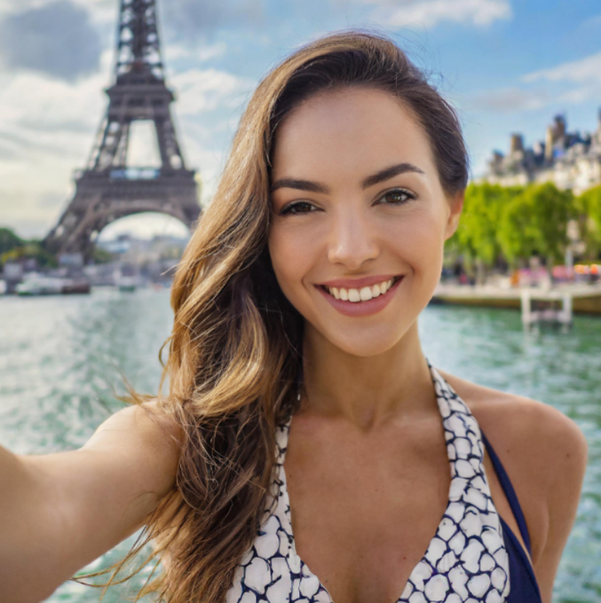 A smiling young woman with long brown hair takes a selfie in front of the Eiffel Tower in Paris, wearing a white and blue patterned halter top.