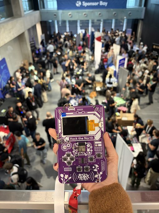 A first-person view of a hand holding a purple circuit board badge labeled 'Hack the North' at a busy indoor event, with a 'Sponsor Bay' banner visible in the background.