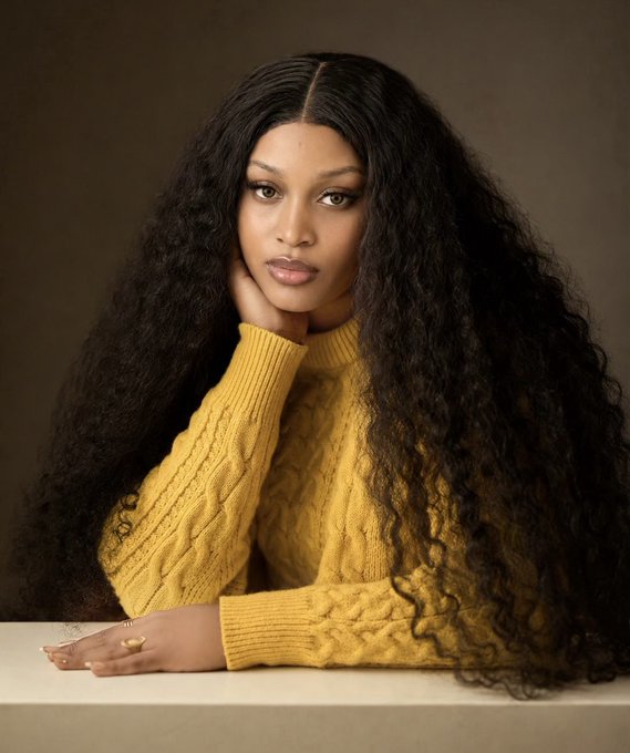 A portrait of a young woman with long, voluminous curly dark hair wearing a yellow cable-knit turtleneck sweater, posing with her hand on her chin.