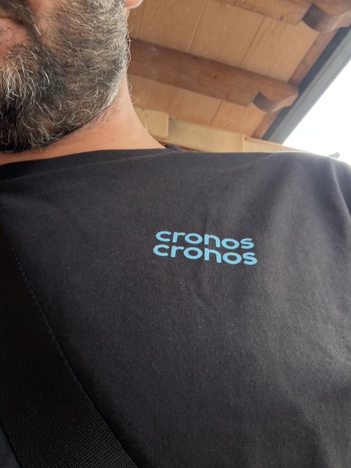A bearded man wears a black t-shirt featuring the 'cronos cronos' logo in blue text.