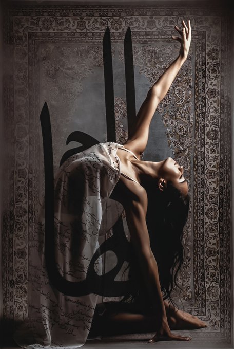 Artistic photograph of a dancer in a flowing sheer dress performing a graceful backbend against an ornate Persian-style carpet backdrop, with dramatic shadow work creating Arabic calligraphy shapes on the floor.