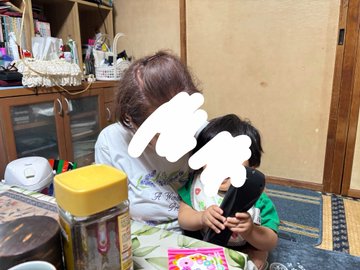 A young woman sits on the floor helping a small child use a phone, with their faces covered by white paint-like edits; the room shows typical home furnishings.