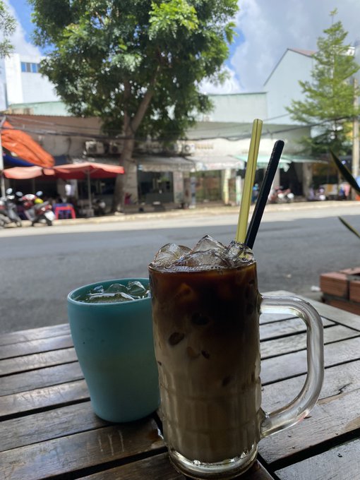 A glass mug of iced coffee with a straw and a blue cup of iced drink sit on a wooden table at a street-side cafe.