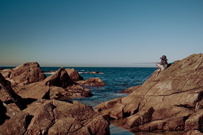 A scenic photograph of a person wearing a hat sitting on a rocky shoreline fishing with a rod, overlooking a calm blue ocean under a clear sky.