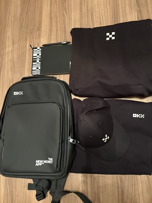 An OKX-branded merchandise bundle on a wooden floor, featuring a black hoodie, backpack, cap, t-shirt, and wallet, with tags reading 'THE NEW MONEY' and 'OKX'.