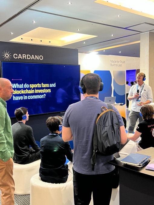 At a Cardano booth, attendees wearing headphones gather around a screen asking 'What do sports fans and blockchain investors have in common?', while a presenter stands in front of a 'Made for Trust Built to Last' display.