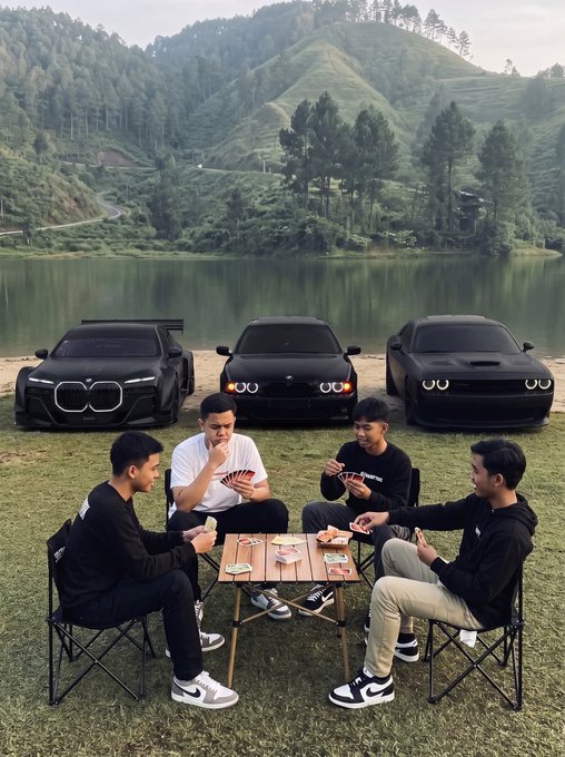 Four young men sit in camping chairs around a small table playing cards, with three black luxury cars (appearing to be BMWs and a Dodge) parked on grass behind them beside a lake and green hills.