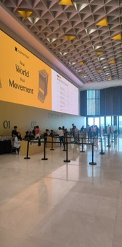 An indoor airport or large hall features a yellow directional sign reading 'World Movement' with the number '01' displayed prominently, alongside queuing stanchions and several people standing around.