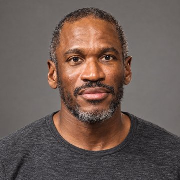 A headshot portrait of an older man with short grey hair and a salt-and-pepper beard, wearing a dark grey t-shirt against a plain grey background.