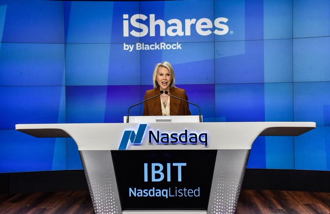 A woman with blonde hair stands at a Nasdaq podium marked 'IBIT NasdaqListed' with an 'iShares by BlackRock' logo displayed on the large screen behind her.