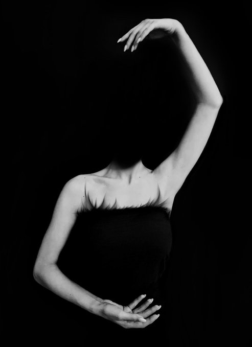 A black and white photograph of a woman in a strapless top posing with her arms raised against a black background.