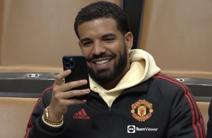 Drake, a known Manchester United supporter, smiles while holding up a phone, wearing a Manchester United jacket with the club crest, Adidas logo, and TeamViewer sponsor clearly visible, tying into his appearance during a Stake.com giveaway tied to the Manchester United vs PSG match.