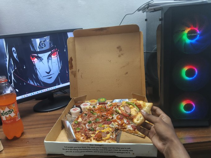 A hand holding a slice of pizza from an open pizza box, with a computer monitor displaying an anime character (Itachi from Naruto), a PC case with RGB lighting, and a bottle of orange soda on a wooden desk.
