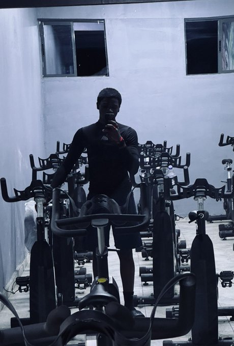 A man stands among stationary exercise bikes in a gym, taking a selfie in a mirror with his phone.