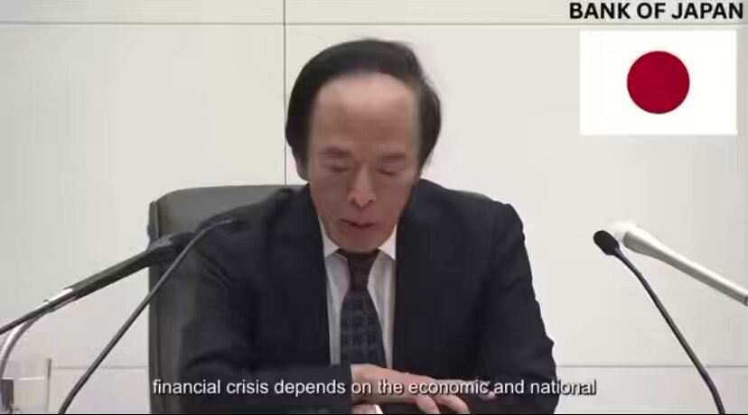 The image shows a man sitting at a desk with two microphones, one of which is placed directly behind him while the other is positioned further back. The man appears to be speaking into the microphone or participating in an interview. The background features a white wall with a red and black flag on it, suggesting that this event may have some connection to Japan's economy or financial matters.
