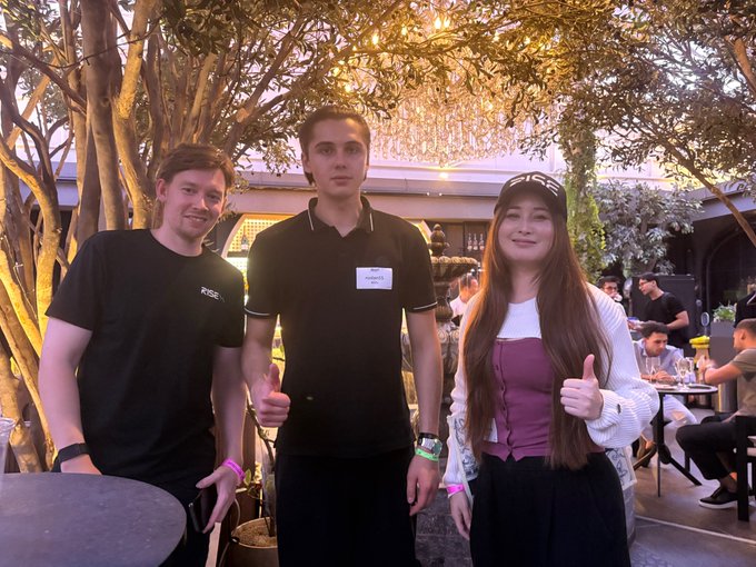 A photograph of three people at an event; a man on the left gives a thumbs up, a taller man in the center wears a name tag, and a woman on the right with a cap also gives a thumbs up.