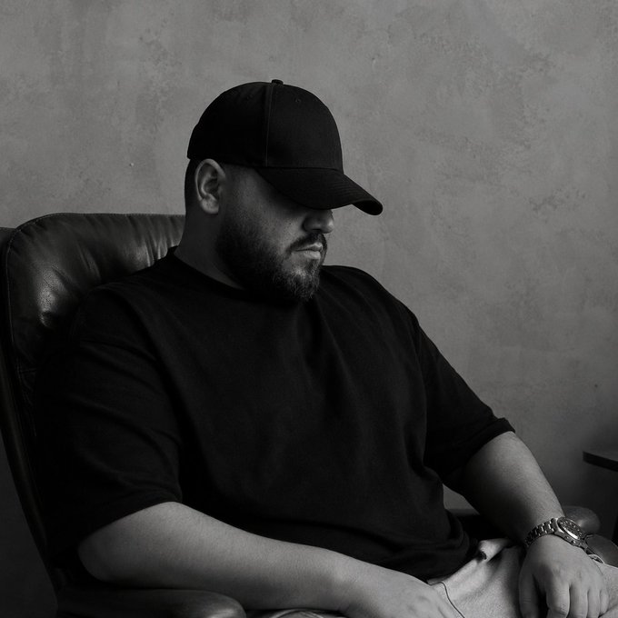 A black and white portrait of a bearded man wearing a black baseball cap and t-shirt, sitting in a leather chair with his head tilted down.