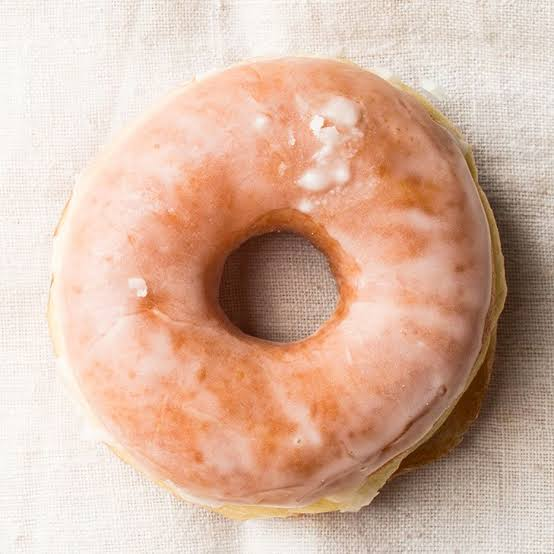 A glazed donut with pink icing resting on a light beige textured fabric surface.