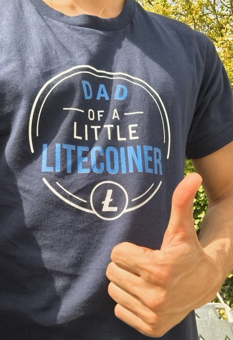 A person wearing a navy t-shirt reading 'DAD OF A LITECOINER' with the Litecoin 'L' logo gives a thumbs-up gesture.