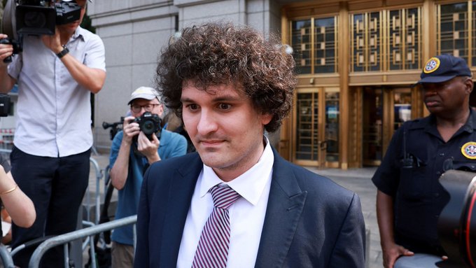Sam Bankman-Fried, the FTX founder with curly hair, exits a building in a dark suit and striped tie as he is surrounded by photographers and a uniformed officer, following the US Senate's unanimous opposition to any potential pardon for him.