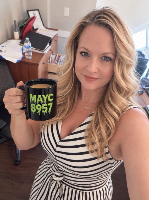 A blonde woman in a black and white striped dress holding up a black mug with green dripping text reading 'MAYC 8957' in what appears to be an office setting.