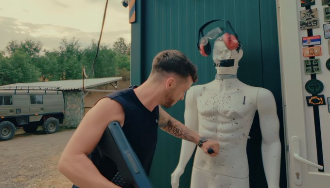 A tattooed man in a sleeveless black shirt holds a laptop and faces a crash test dummy wearing ear protection, standing outside near a vintage off-road vehicle and a shipping container. The scene suggests training or preparation, fitting the post's reference to being "ready for Breakpoint, London," likely indicating a cybersecurity conference.