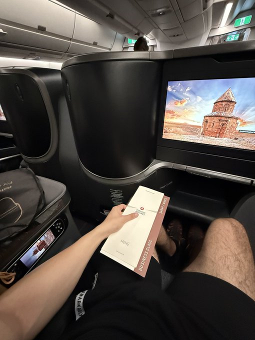 A traveler settles into a business class seat, holding a "MENU" labeled "BUSINESS CLASS" while a screen displays an image of a church, capturing the anticipation of heading to Coinfest and exploring events like MEXC and Alpha Arena.