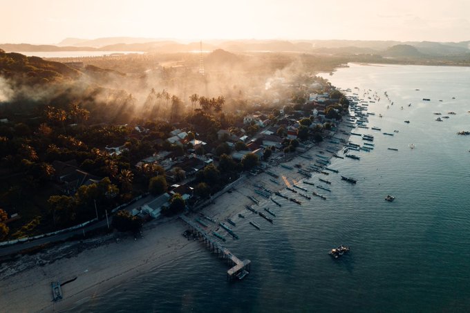 An aerial view captures a coastal town with boats lined up along the shore at sunset, with mist rising over the trees and hills in the background.