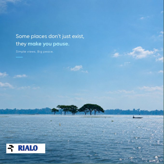 A scenic view of a wide, calm body of water under a blue sky with scattered clouds, featuring a small island with trees and a tiny boat. Text overlay reads: 'Some places don't just exist, they make you pause. Simple views. Big peace.' with a 'RIALO' logo in the bottom left.