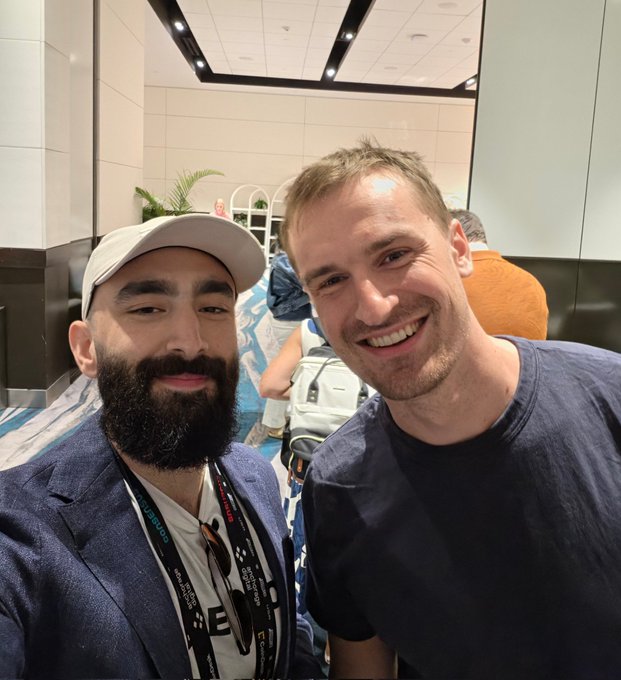 The image shows two men posing for a picture with smiles on their faces. One of them has a beard and is wearing a blue jacket. The other man, who appears to be balding, is also smiling at the camera. They are standing in front of a white wall that seems to have a window behind it. There is another person partially visible in the background, but they don't seem to be the main focus of the photo.