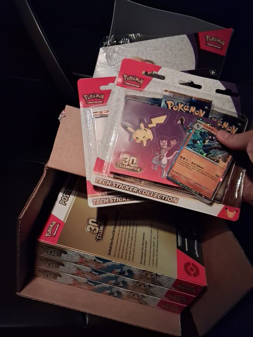 A hand holds up a Pokémon 30th Celebration Tech Sticker Collection pack and a holographic Pikachu promo card, pulled from a box stacked with multiple Pokémon trading card game products. The celebratory haul lines up perfectly with the "Bag secured!!!" caption, capturing the excitement of unboxing a successful pickup.